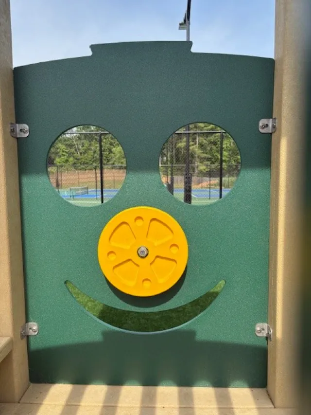 Green playground panel with two circular windows, a yellow wheel, and a curved cutout resembling a smiling face.