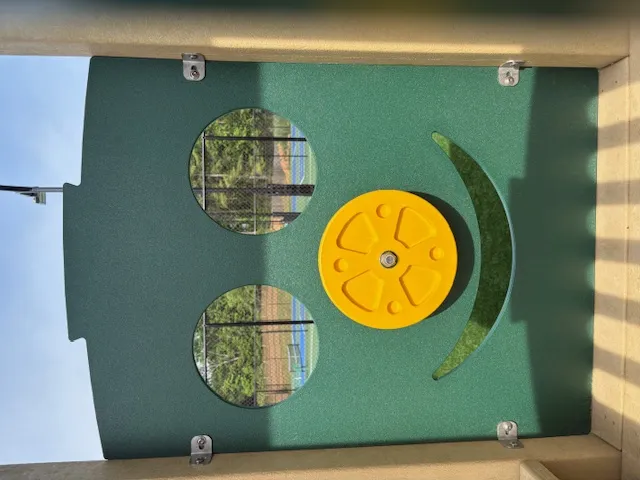 Green playground panel with two circular windows, a yellow wheel, and a curved cutout resembling a smiling face.