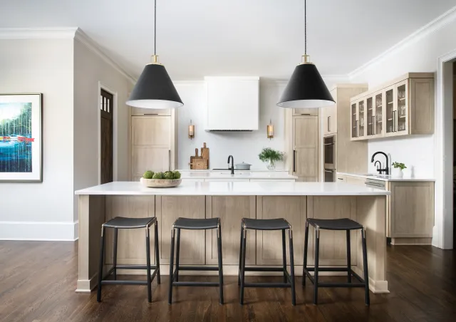 Modern kitchen with a large island, four black stools, pendant lights, and wood flooring under natural light.