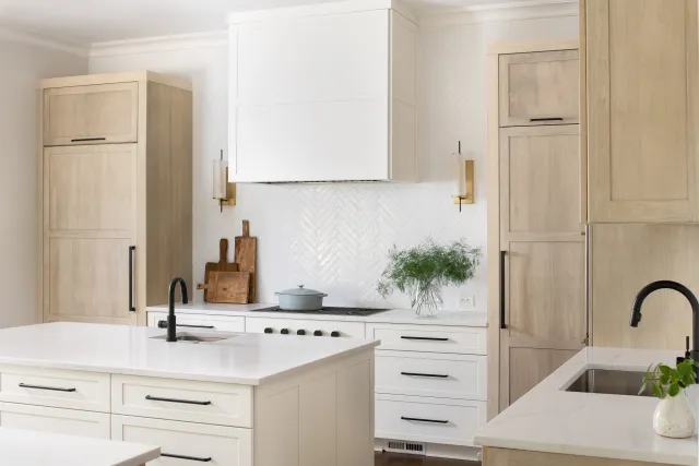 Modern white kitchen with wooden cabinets, marble countertops, dual black faucets, and green plant decoration.