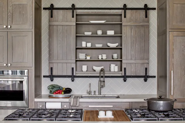 Modern kitchen with gray wood cabinets, sliding barn doors, gas stove, and open shelves holding white dishware.