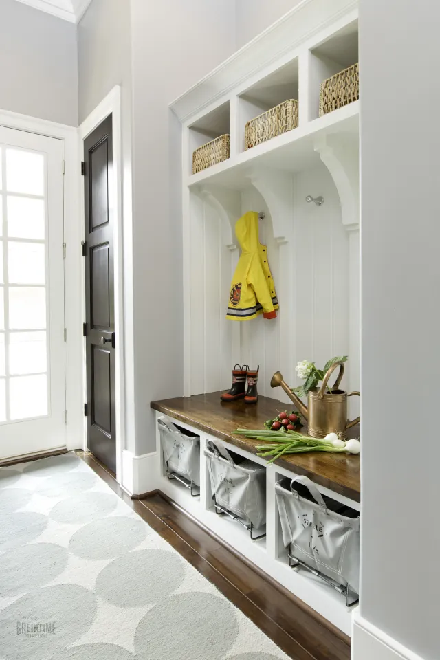 Organized mudroom with white cubbies, wooden bench, yellow raincoat, boots, and gardening supplies on a patterned rug.