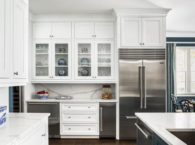 Modern white kitchen with glass-front cabinets, stainless steel appliances, marble countertops, and blue decorative vases.