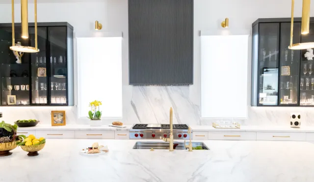 Bright modern kitchen with marble countertops, gold fixtures, black cabinets, and fresh fruit bowls