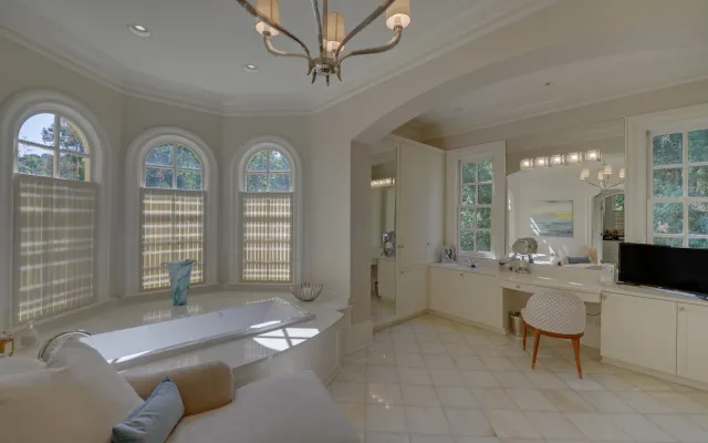 Bright luxurious bathroom with a large bathtub, arched windows, chandelier, and built-in vanity with seating.