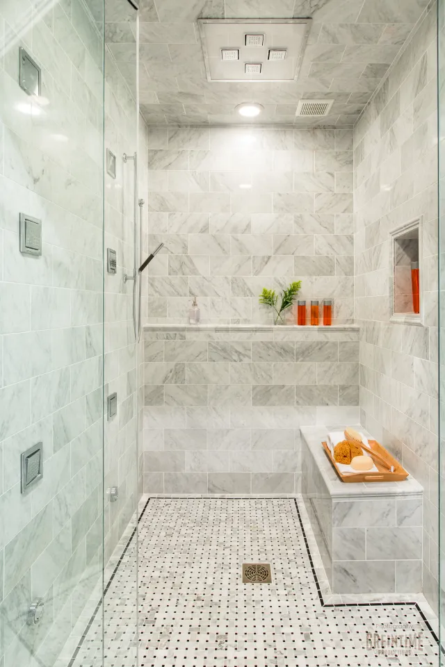 Modern walk-in shower with marble tiles, glass door, bench with bath accessories, and rainfall showerhead.