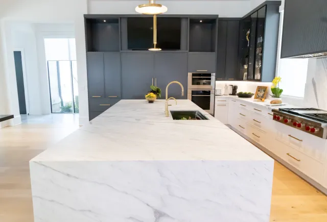 Modern kitchen with large white marble island, black cabinets, gold fixtures, and stainless steel appliances.