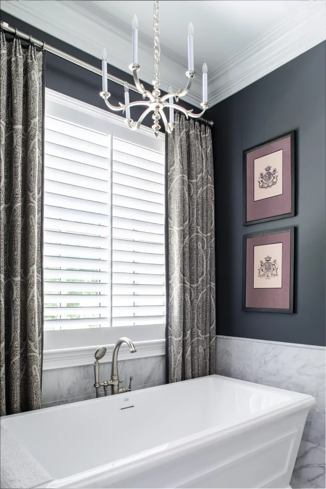 Modern bathroom with white bathtub, silver faucet, patterned curtains, chandelier, and framed wall art.