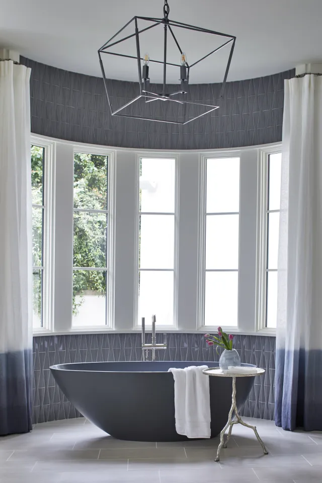 Modern bathroom with a black freestanding bathtub, large windows, white curtains, and geometric tile walls.