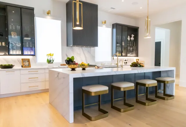 Modern kitchen with large marble island, four bar stools, dark cabinetry, and gold pendant lights over hardwood floor.