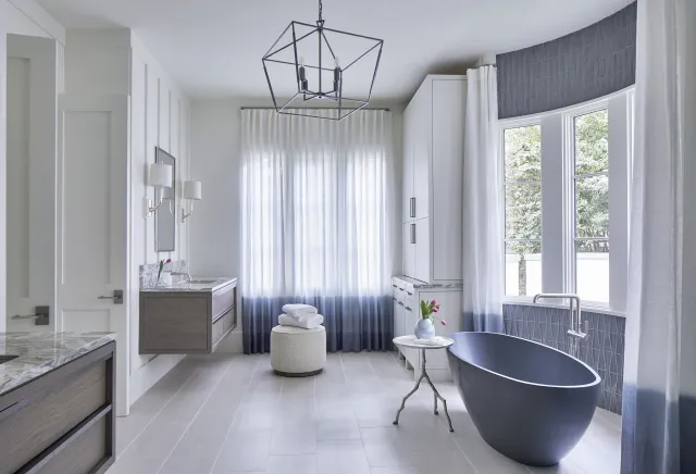 Modern bathroom with freestanding black tub, large windows, gray floor tiles, floating vanities, and sheer gradient curtains.