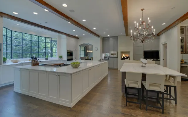 Spacious modern kitchen with large white islands, wooden beams, chandelier, and hardwood flooring under natural light.