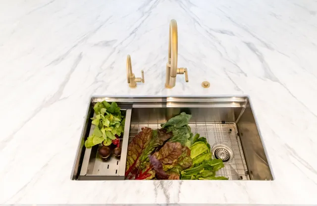 Overhead view of stainless steel kitchen sink with fresh leafy greens and vegetables on white marble countertop.