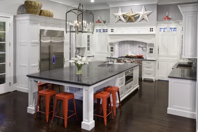 Modern white kitchen with large black granite island, orange stools, stainless steel appliances, and star wall decor.