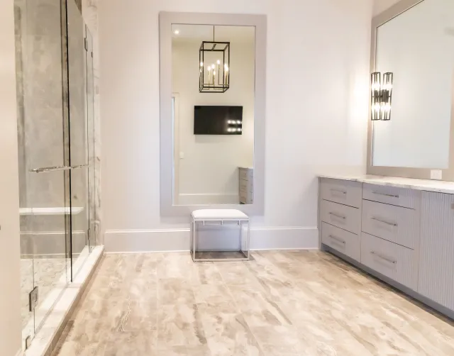 Bright modern bathroom with large mirror, glass shower door, light fixture, and gray cabinetry with marble floor.