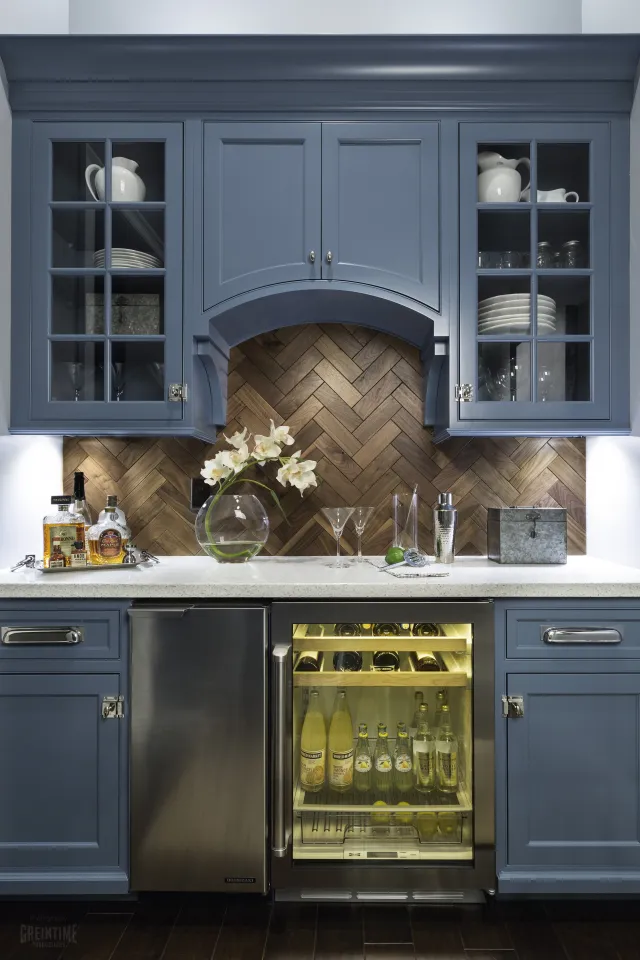 Modern kitchen bar area with blue cabinets, wood herringbone backsplash, and countertop drinks fridge.