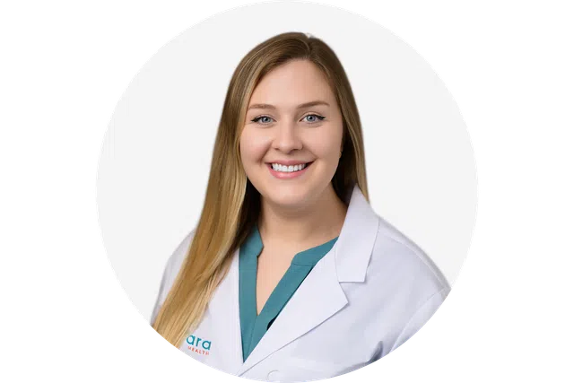 Smiling female healthcare professional with long blonde hair wearing a white lab coat and teal scrubs