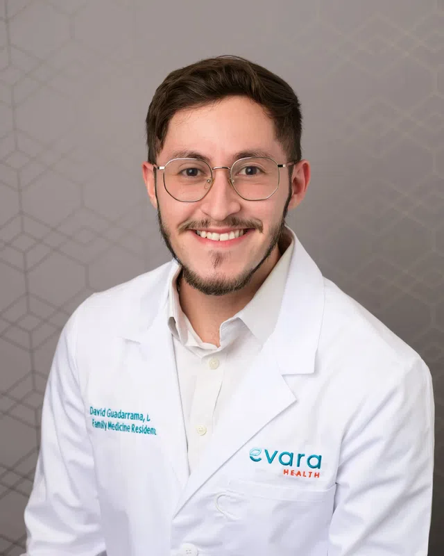 Smiling male doctor wearing glasses and a white coat with Evara Health logo and family medicine resident title.
