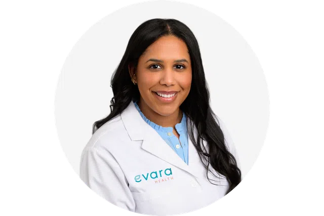 Smiling female healthcare professional wearing Evara Health white coat and blue blouse with long hair