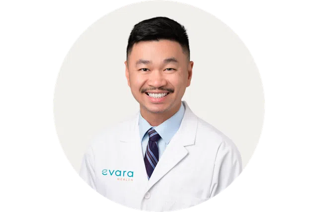 Smiling male doctor wearing a white Evara Health lab coat and striped tie against a patterned background