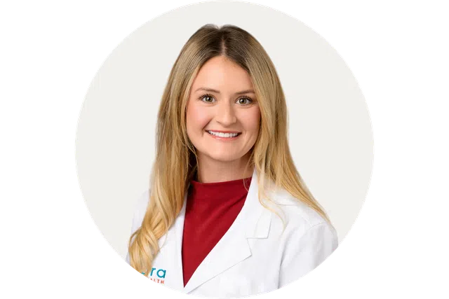 Smiling female doctor with long blonde hair wearing a white coat and red top on a white background.