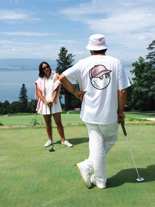 Two people dressed in white golfing attire playing on a green golf course overlooking a lake and trees under a blue sky.