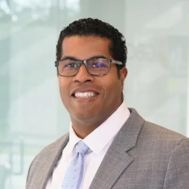 Smiling man wearing glasses, gray checkered suit, white shirt, and light blue polka dot tie in a bright office.