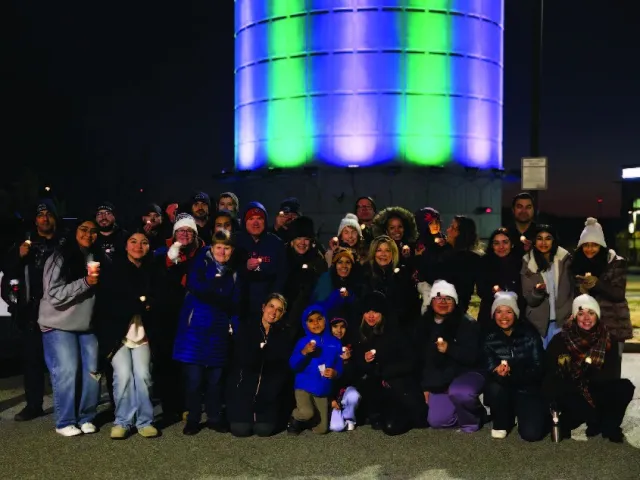 Group of people gathered at night holding candles in front of a tower lit with green and blue lights.