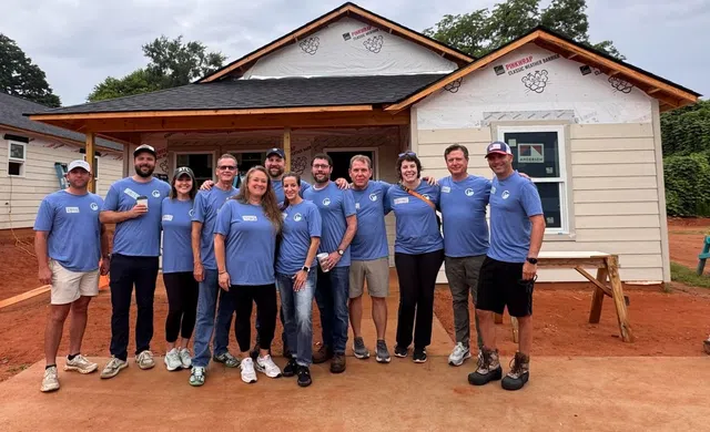 Group of volunteers in matching blue shirts posing in front of a newly constructed house under cloudy sky.