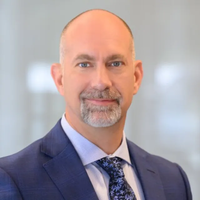Professional middle-aged man in a blue suit and patterned tie smiling against a blurred office background