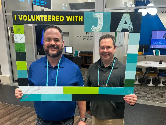 Two adults read books to young children seated on a colorful classroom carpet with educational posters.