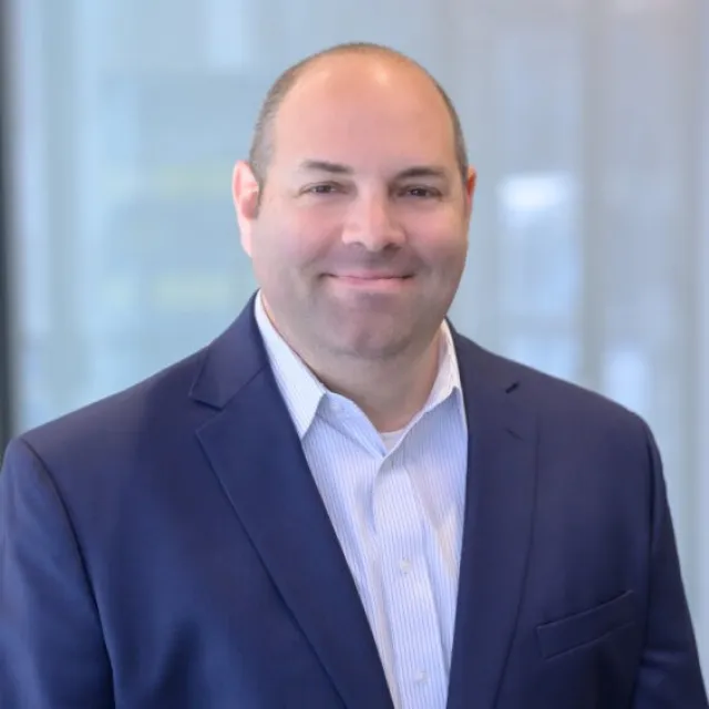 Professional man in a blue suit jacket and white shirt smiling with a blurred office background.