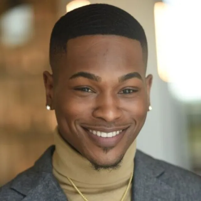 Smiling man wearing a tan turtleneck, gray blazer, gold cross necklace, and diamond earrings in soft lighting.