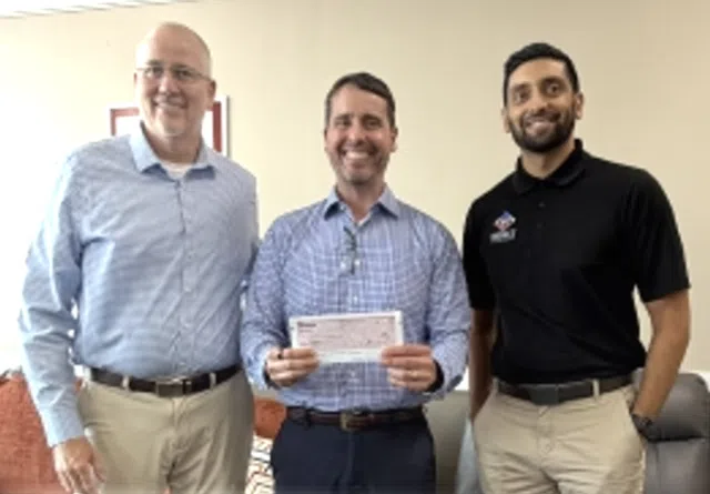 Three men standing indoors smiling, middle man holding a check, casual business attire, friendly atmosphere.