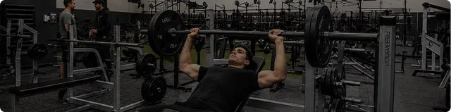 Man lifting heavy barbell on bench press in a gym filled with workout equipment and people training.