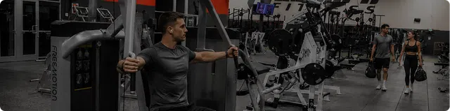 Man using strength training machine in a modern gym with various fitness equipment and two people walking in background
