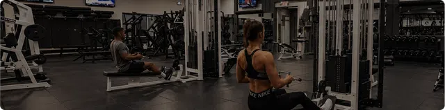 Two people exercising on rowing machines in a spacious, modern gym with weight equipment visible.