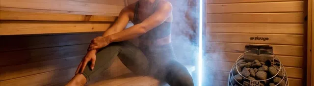 Woman relaxing in a wooden sauna surrounded by steam, wearing workout clothes and sitting on a bench.