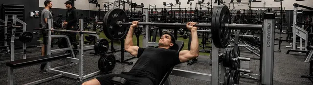 Athlete performs bench press in a modern gym with fitness equipment and TV screens showing sports and news.