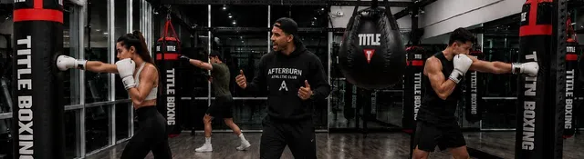 Boxing coach instructs female and male trainees punching heavy bags in a spacious gym with mirrors and wooden floor.
