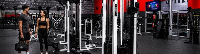 Modern gym interior with weight machines, fitness equipment, and two people carrying gym bags inside.