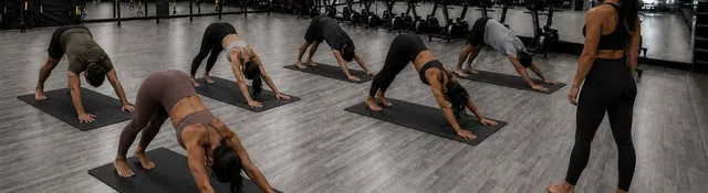 Group yoga class in downward dog pose led by instructor in modern gym studio with wooden floors and mirrors.