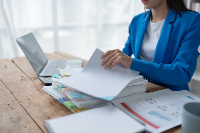 Professional woman in blue blazer organizing large stack of documents at wooden desk with laptop and charts.