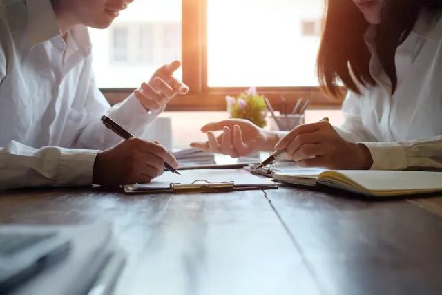 Two professionals discussing documents and writing notes at a wooden table by a sunlit window