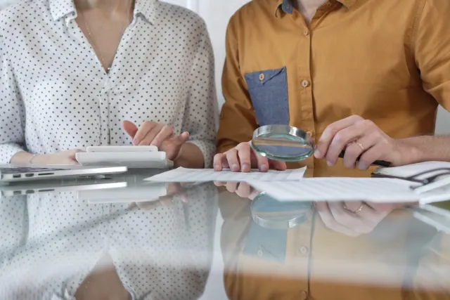 Two people reviewing documents with a magnifying glass and calculator on a reflective table surface.