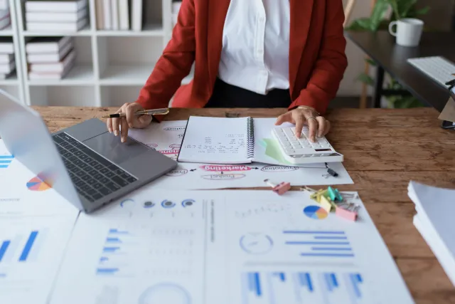 Person in red blazer working with calculator, laptop, and financial charts on a wooden desk in office.