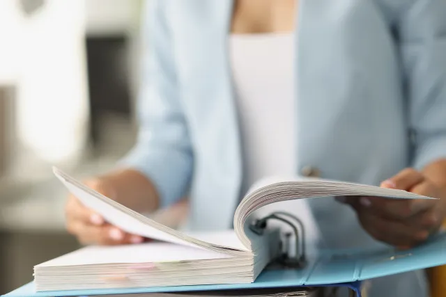 Person in blue jacket flipping through a thick binder full of documents on a desk.
