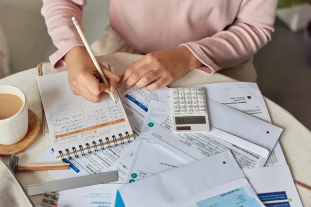 Person calculating and writing monthly bills on notepad surrounded by receipts, calculator, and coffee on table.
