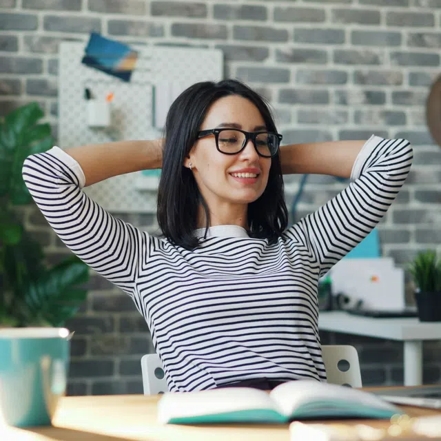Woman in glasses relaxing at desk with laptop, book, and coffee cup in modern office space