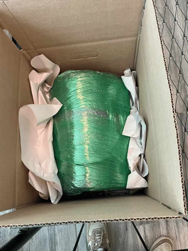 A green wrapped item cushioned with white packing paper inside a cardboard box on a wooden floor with a shoe visible.